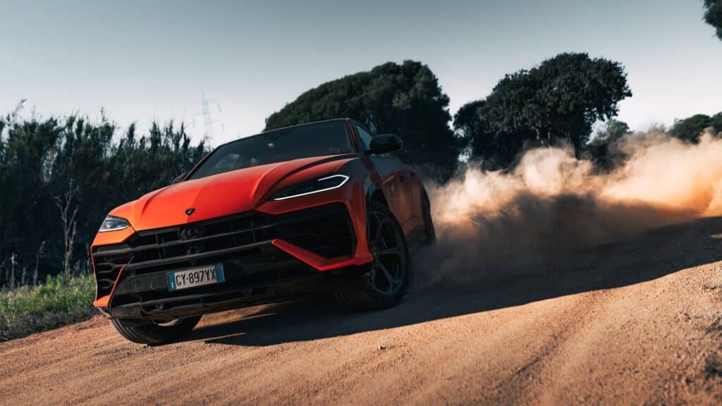 Les 10 SUV de luxe les plus sportifs en 2026 : Lamborghini Urus SE orange vue de trois-quarts avant sur une piste de terre, soulevant un nuage